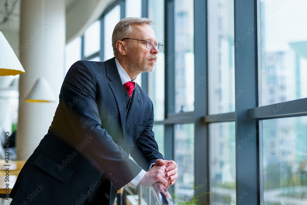 Thoughtful senior businessman looking through window with his hands on chair. Thoughtful senior businessman looking through window with his hands on chair.