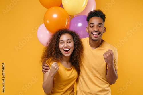 Happy young friends couple african american guy girl in casual clothes isolated on yellow background. Birthday holiday party concept. Celebrate hold colorful air balloons hugging doing winner gesture. © ViDi Studio