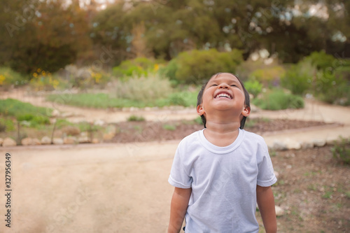 A little boy with a white tshirt is expressing joy as he looks to the sky and laughs, standing at the end of a dirt path or trail.