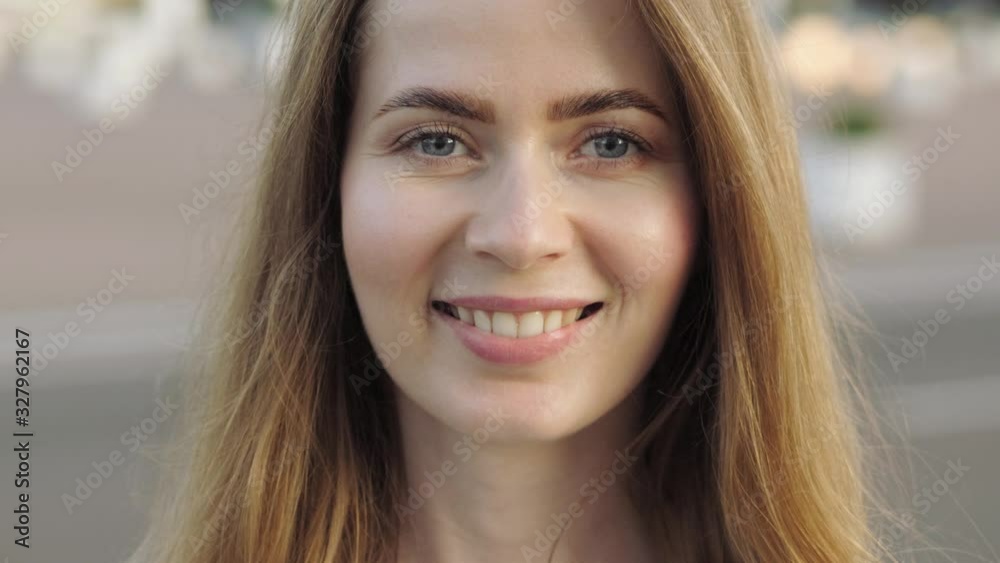 Big close up slow motion head shot of young adorable blonde elegant woman with blue eyes looking at the camera changing facial expression from serious to adorable smile on city street golden hour warm