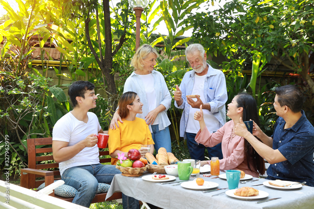 Happy multiethnic family standing and sitting at a breakfast table in backyard outdoor on sunny day with smiling face. Happy caucasian and asian family sitting and having conversation with good time. Happy multiethnic family standing and sitting at a breakfast table in backyard outdoor on sunny day with smiling face. Happy caucasian and asian family sitting and having conversation with good time.