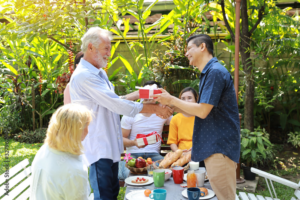 Obraz premium Happy multiethnic family giving surprise gift to caucasian grandfather on his happy birthday with happy smiling face in backyard outdoor on sunny day. Elder men shaking hands outdoor with happy face.