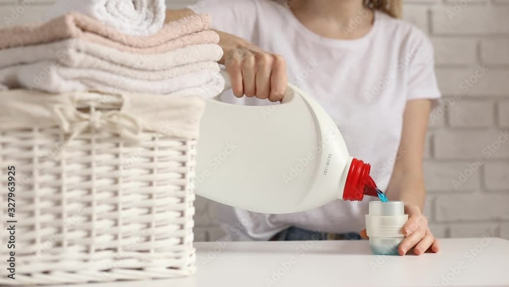 Close up of female hands pouring liquid laundry detergent into cap on white rustic table with towels in wicker basket