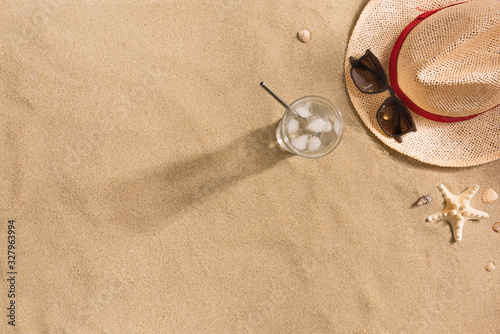 Beautiful summer holiday beach background with straw hat, sunglasses, glass of water and shells on sand background, top view with copy space. Harsh shadows