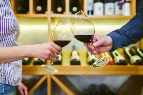 Close up front view on male and female caucasian hands holding glasses of red wine toasting in front of the shelf man and woman celebrating concept