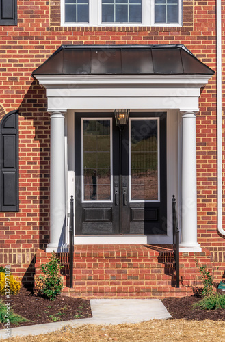 View of classic portico entrance covered w/ dark color metal roof, supported by two colonial white vinyl wrap columns w/ decorative caps and trim black baluster railing on a brick luxury American home