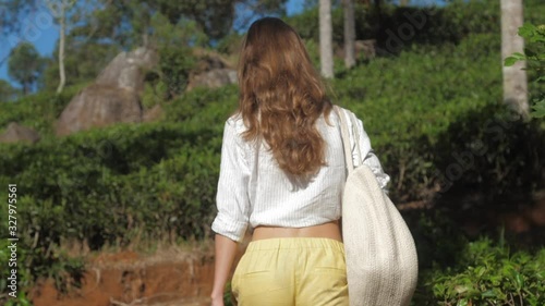 blonde long haired girl with large handbag walks in tropical park past slope backside view slow motion. Concept ecology environment