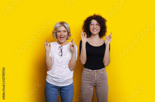 Cheerful caucasian girl with curly hair is dreaming about something near her blonde friend while crossing the fingers on a yellow wall