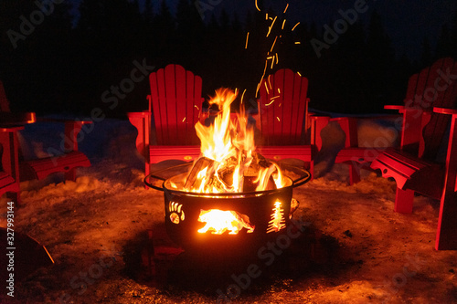 Cozy/romantic evening scene of warming bonfire on snow in winter with red chairs around. Canadian symbols of bear, paw and tree on the fire container. Banff National Park, Alberta, Canada