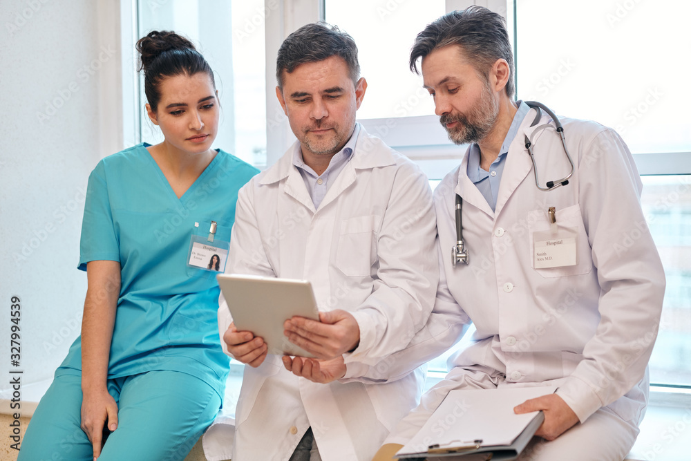 Group of medical specialists sitting on window sill and using tablet while discussing online patients card Group of medical specialists sitting on window sill and using tablet while discussing online patients card