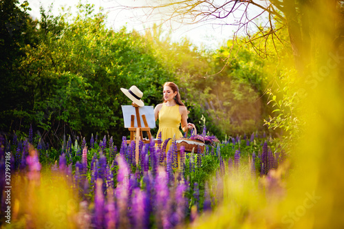 A girl artist in a yellow dress and hat draws a lupine field in the open air. Easel box with canvas, oil paint, palette and artist 's tools. © Elena 