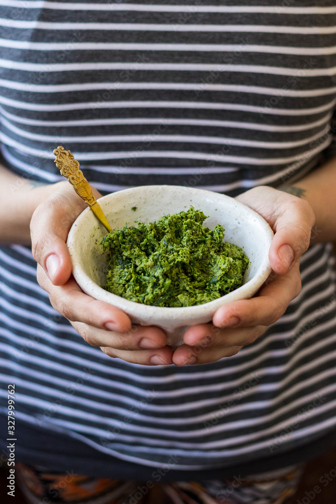Green basil pesto paste dip sauce  in bowl with seeds, nuts and green herbs. Hands hold pesto.