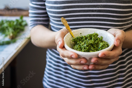 Green basil pesto paste dip sauce in bowl with seeds, nuts and green herbs. Hands hold pesto.