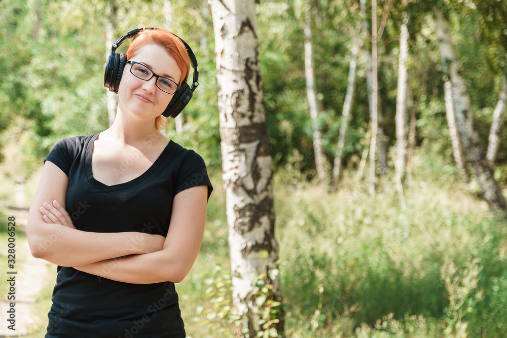 Plump ginger girl in in glasses and headphones posing in summer birch forest. Music and body positive concept, copy space. Plump ginger girl in in glasses and headphones posing in summer birch forest. Music and body positive concept, copy space.