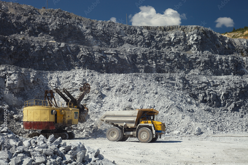 Obraz premium Mining dump truck drives off from the excavator after loading in a quarry for the extraction of limestone on the background of rocky terrain and blue sky in sunny weather.