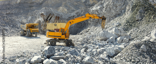 Hydraulic breaker hammer in a quarry for limestone mining with an excavator and a quarry truck in the background, panorama.