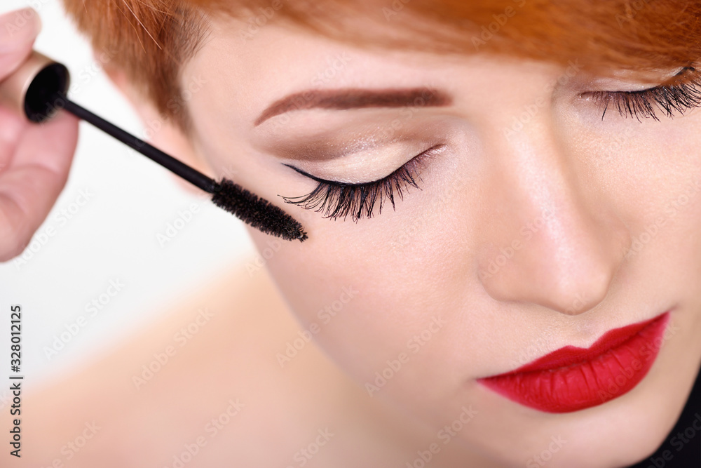 Makeup artist applies mascara brush. Beautiful young woman with short red hair. Makeup detail.