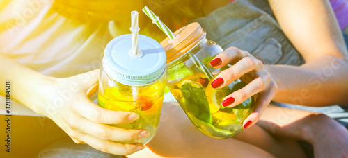 Friends holding fresh lemonade in jars with straws. Hipster summer party with drinks. Healthy vegan lifestyle. Eco-friendly in the nature. Lemons, oranges and berries with mint in the glass.