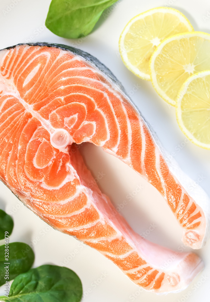 A piece of fresh salmon fish steak on a white plate with spinach and lemon, on a gray wooden background. Omega 3 vitamin, healthy lifestyle. Natural vegetarian food. Top view. A piece of fresh salmon fish steak on a white plate with spinach and lemon, on a gray wooden background. Omega 3 vitamin, healthy lifestyle. Natural vegetarian food. Top view.