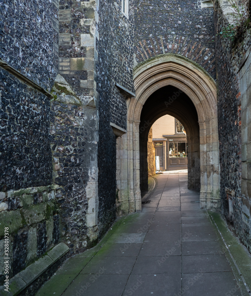 Dark and narrow medieval alleyway with flint walls either side and an architectural arch at the end