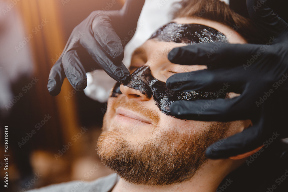 Barber applies black charcoal mask to man face to clean pore skin and remove acne from nose, spa salon