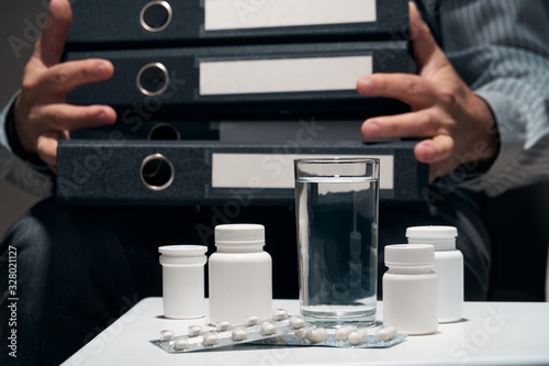 Overworked businessman sitting on the sofa and taking medication. Pills and glass of water near him.