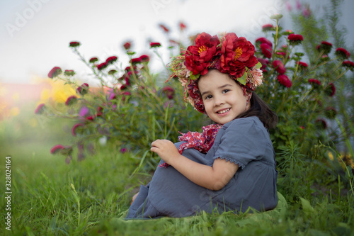 A little girl with a red floral crown sits under a flowering bush with burgundy carnations