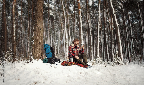 Outdoorsman relaxing drinking tea from thermos sitting on traveling rug at forest edge in winter
