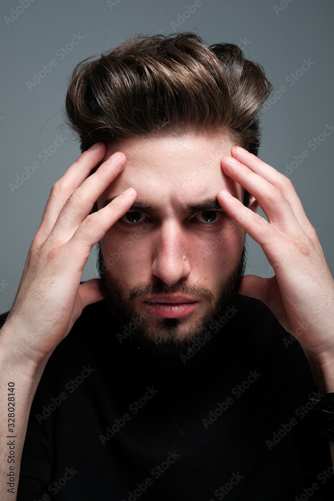 A young dark-haired man with a beard holds his head and shows different human emotions: hatred, fear, despair, horror, malaise, headache, clairvoyance. Close-up studio portrait of a man.