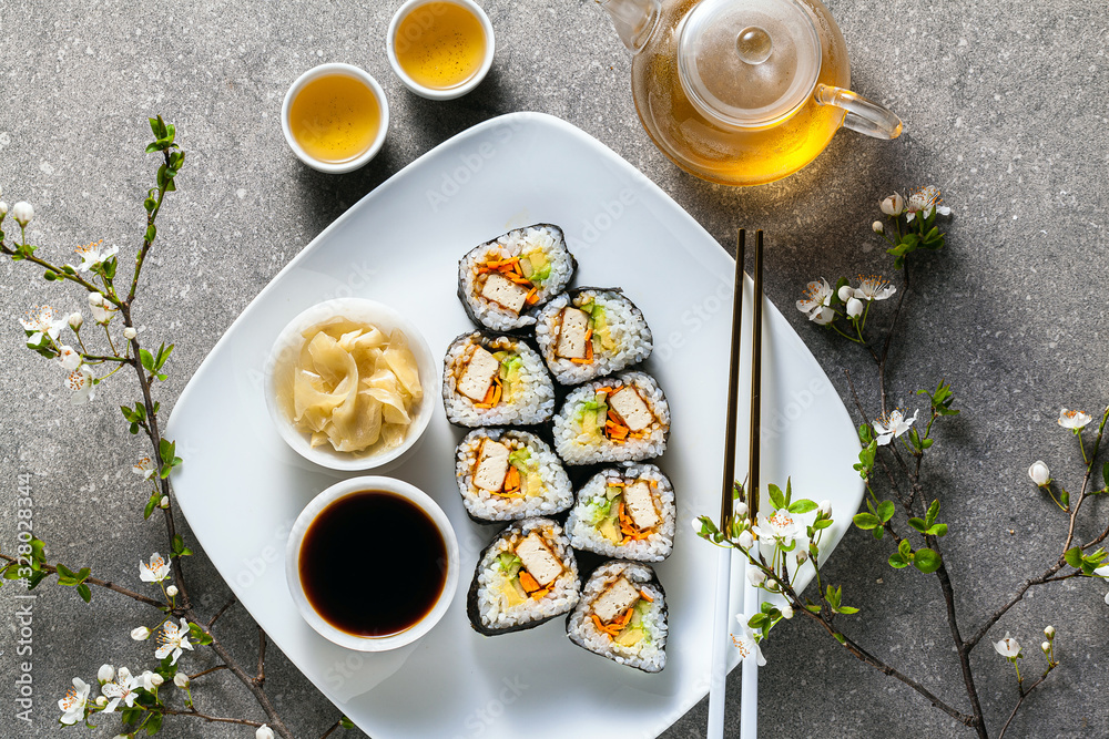 vegan sushi with tofu in General Tso sauce, avocado and fresh vegetables . on a table with branches of a blossoming tree and green tea in a glass teapot
