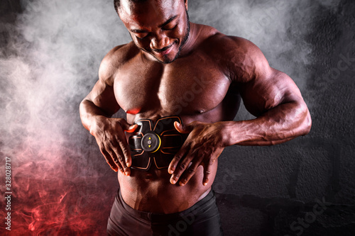 Bodybuilder male African uses electronic belt muscle stimulator trainer abdominal muscles on a black background with smoke