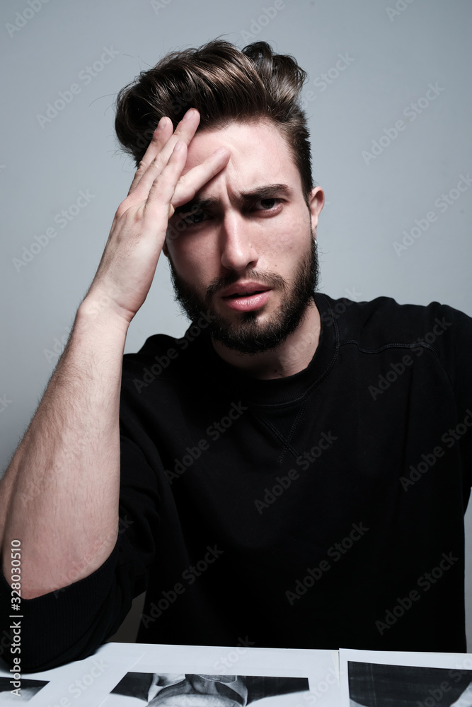 A young dark-haired man with a beard holds his head and shows different human emotions: hatred, fear, despair, horror, malaise, headache, clairvoyance. Close-up studio portrait of a man. A young dark-haired man with a beard holds his head and shows different human emotions: hatred, fear, despair, horror, malaise, headache, clairvoyance. Close-up studio portrait of a man.
