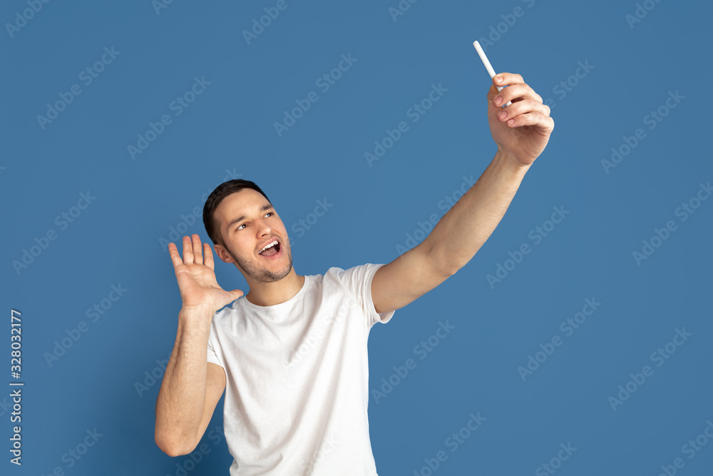 Taking selfie. Caucasian young man's portrait isolated on blue studio background. Beautiful male model in casual style, pastel colors. Concept of human emotions, facial expression, sales, ad. Taking selfie. Caucasian young man's portrait isolated on blue studio background. Beautiful male model in casual style, pastel colors. Concept of human emotions, facial expression, sales, ad.