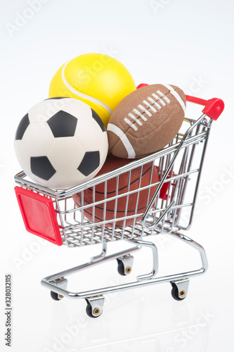 Shopping cart on white background filled with sporting goods including soccer ball, American football, tennis ball, and basketball