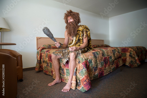 Bored caveman sitting indoors on the edge of a bed with a colorful bedspread in an anonymous cheap motel room