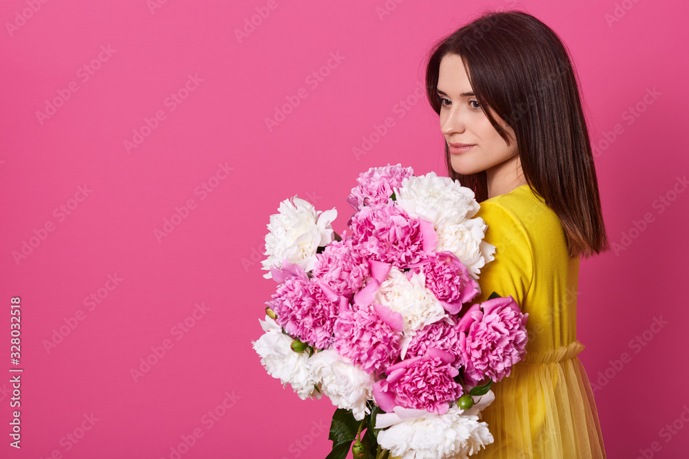 Φωτογραφία Studio shot of charming woman wearing yellow dress isolated over rosy studio background, holding white and pink peony flowers, looking at aside, looks romantic and gentle