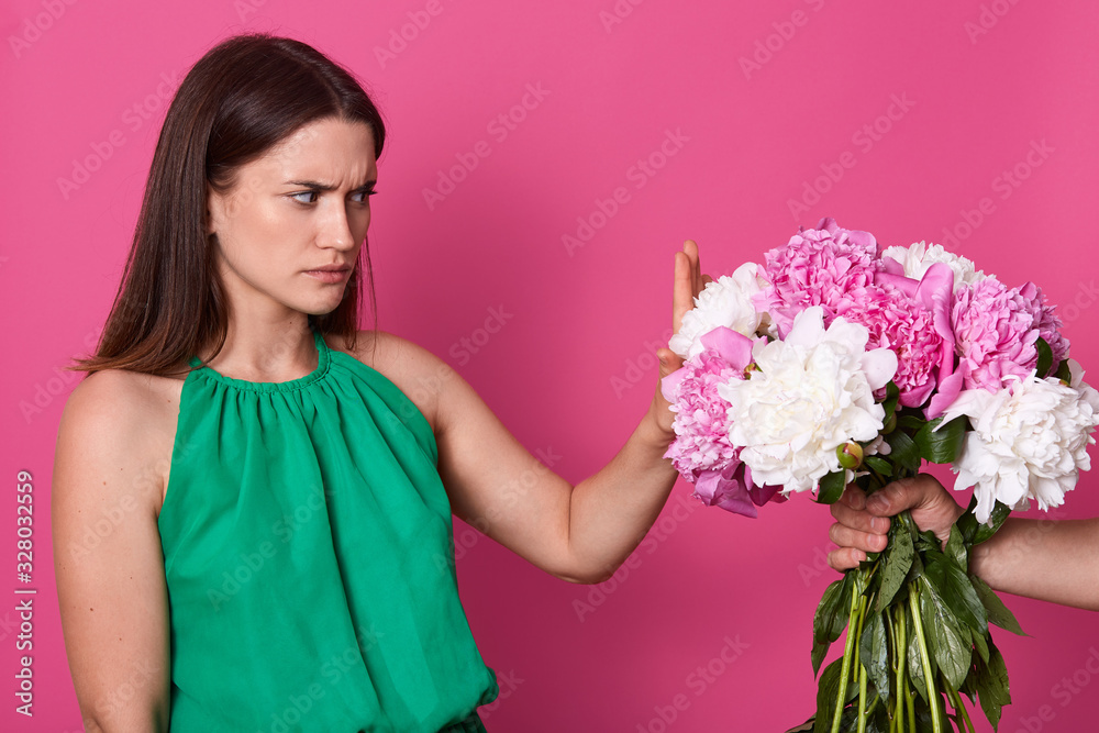 Indoor shot of attractive lady dressed green sundress, refuses beautiful bouquet of white and pink peony flowers from faceless person, brunette girl with offended facial expression, against rose wall. Indoor shot of attractive lady dressed green sundress, refuses beautiful bouquet of white and pink peony flowers from faceless person, brunette girl with offended facial expression, against rose wall.