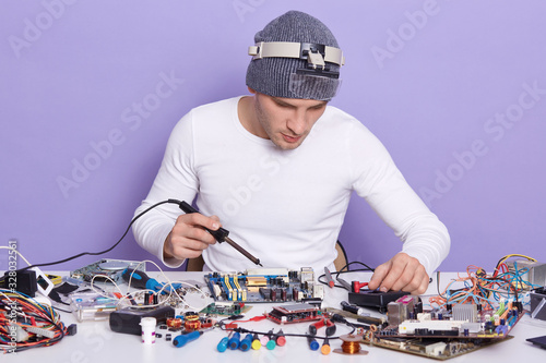 Close up portrait of young PC technician working with soldering iron and repairing computer motherboard isolated over lilac background, radioman dresses white shirt and gray cap, looks concentrated. photo