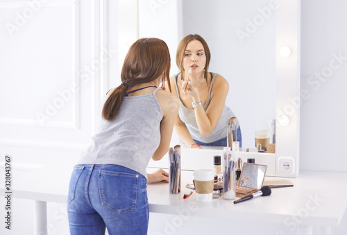 Amazing young woman doing her makeup in front of mirror. Portrait of beautiful girl near cosmetic table.