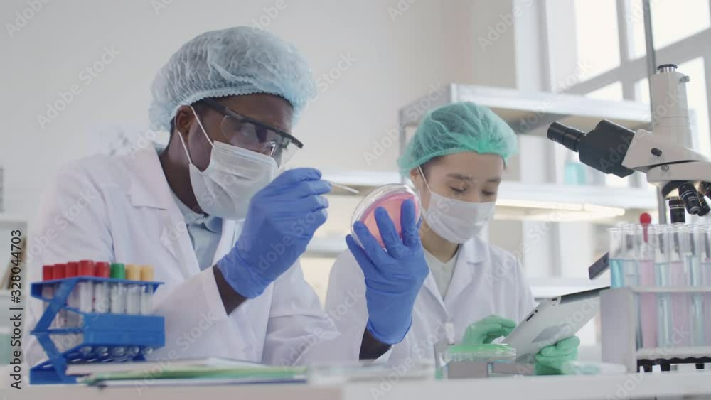 Medium shot of African man and Asian woman wearing special uniform sitting together in lab and working