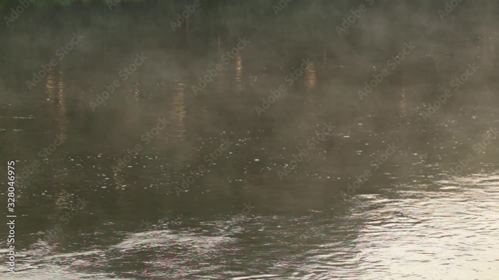 Morning white fog moving over calm surface of countyrside river.