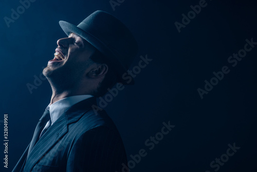 Mafioso in suit and felt hat laughing on dark background