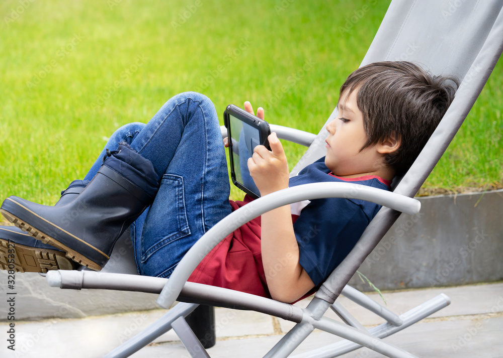 Obraz premium Portrait kid with playing game on tablet, A boy having fun watching cartoons on digital taplet, Child siting on roacking chair relaxing on weekend in the garden on spring or Summer