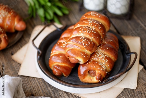 Homemade sausages in yeast dough with sesame seeds and milk on a wooden background.