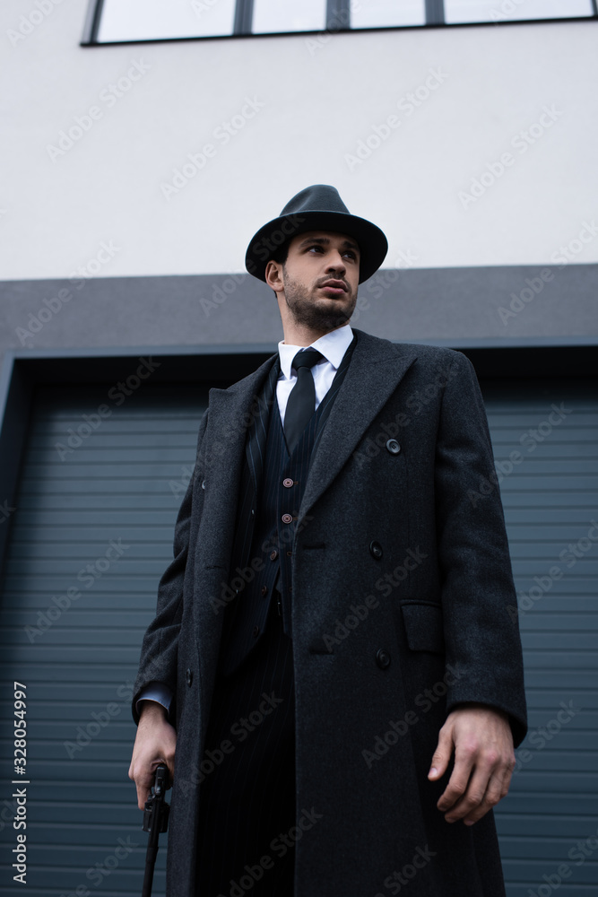 Fototapeta premium Low angle view of gangster in black coat and hat with weapon on street