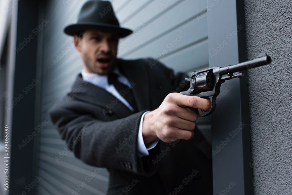 Selective focus of mafioso aiming gun with outstretched hand near wall on street Selective focus of mafioso aiming gun with outstretched hand near wall on street