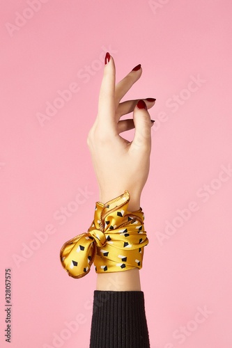 Close-up shot of a female hand wrapped with a yellow silk handkerchief with black and white all-over geometric pattern. The photo is made on the pink background.   photo