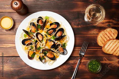 Mussels and pasta with bisque sauce, lie on a white plate. Next to it is parsley sauce, a glass of wine, white fried bread and a fork. View from above