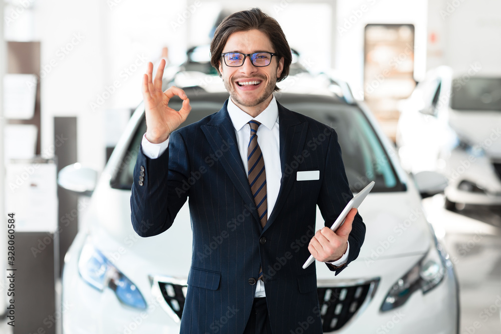 Smiling Salesman Gesturing Okay Standing In Luxury Auto Store Smiling Salesman Gesturing Okay Standing In Luxury Auto Store