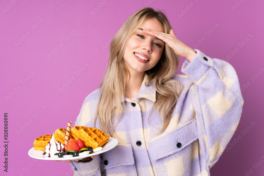 Teenager Russian girl holding waffles isolated on purple background looking far away with hand to look something Teenager Russian girl holding waffles isolated on purple background looking far away with hand to look something